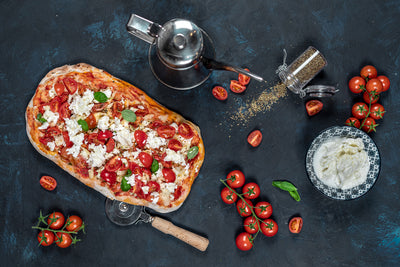 Roman-Style Buffalo Mozzarella Cherry Tomatoes Pinsa with basil on dark blue backdrop, pizza cutter, and fresh ingredients on the side